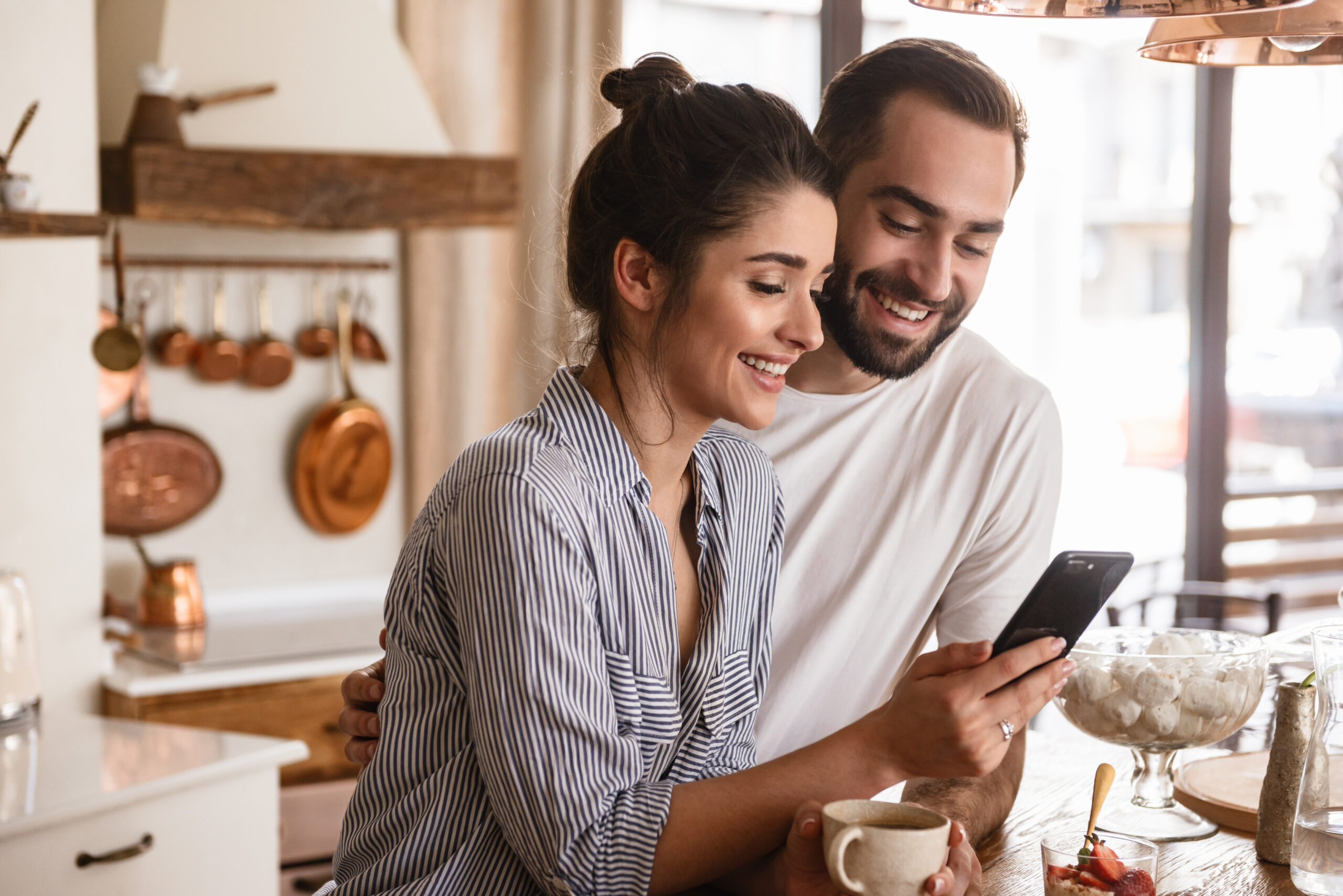 Photo of smiling brunette couple man and woman 20s drinking coffee and using cell phone during breakfast in kitchen at home