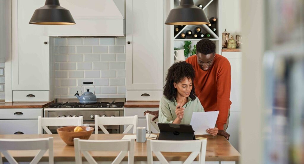Young multiracial couple smiling while going over bills and doing online banking on a digital tablet in their kitchen at home