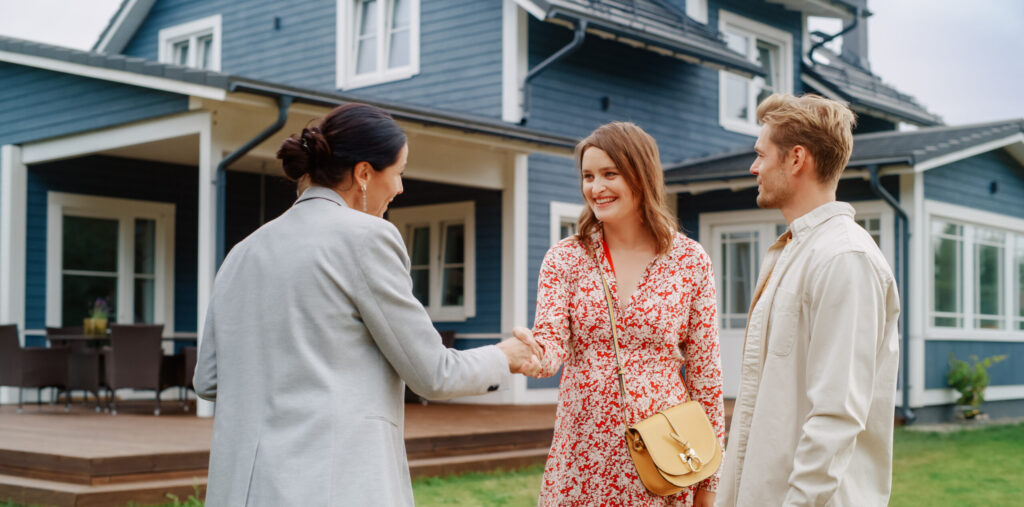 Happy Couple Shaking Hands with Realtor in Front of Their New Home. Professional Real Estate Agent Congratulating Young Homeowners with Property Purchase, Giving Them the Key to the House.