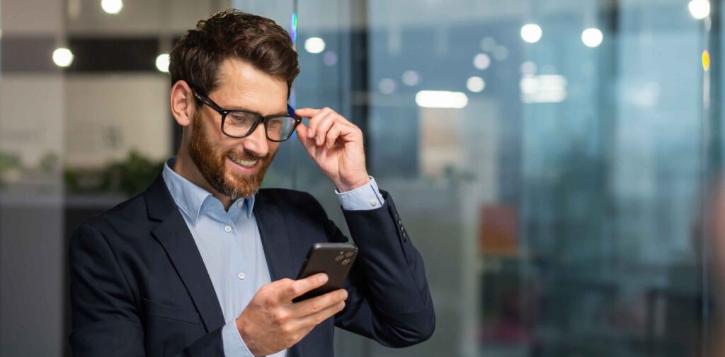 Successful financier investor works inside office at work, businessman in business suit uses telephone near window, man smiles and reads good news online from smartphone.