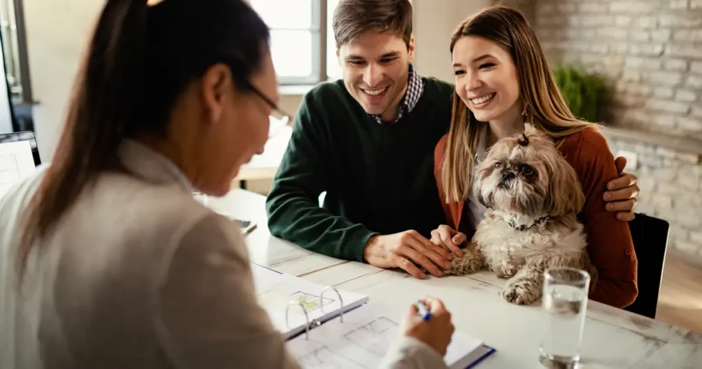 A smiling couple sits across from a mortgage advisor at a desk, discussing home loan options, with their small fluffy dog on their lap.