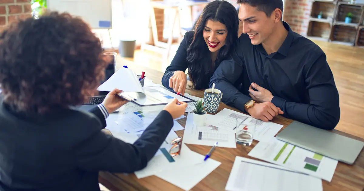 A smiling couple in business attire reviews financial documents with a mortgage advisor in a modern office, discussing home loan options.