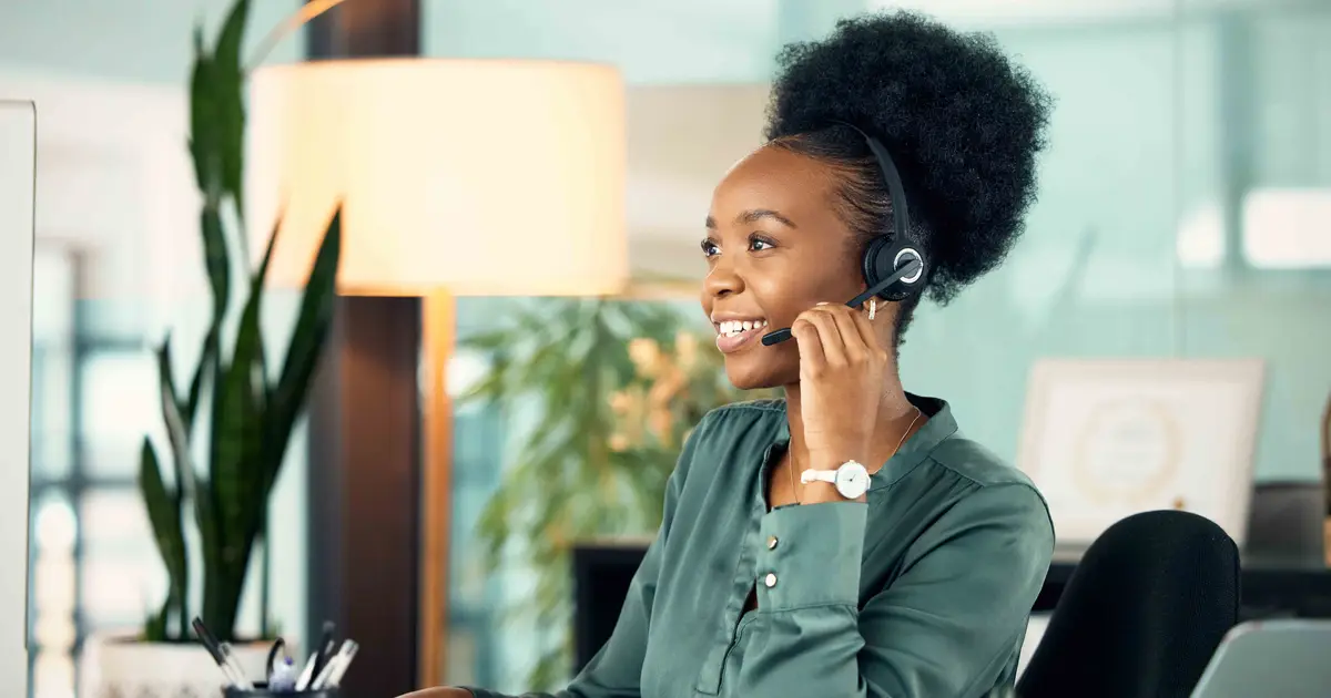 A smiling customer service representative wearing a headset sits at a desk in a modern office, engaged in a phone conversation.