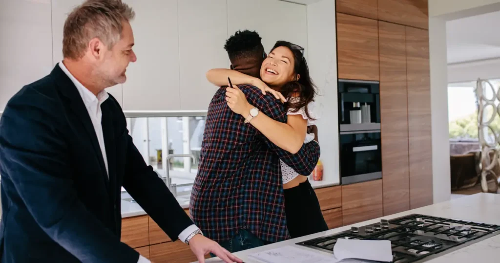 A happy couple embraces in a modern kitchen after signing home purchase documents, while a real estate agent smiles and prepares paperwork.