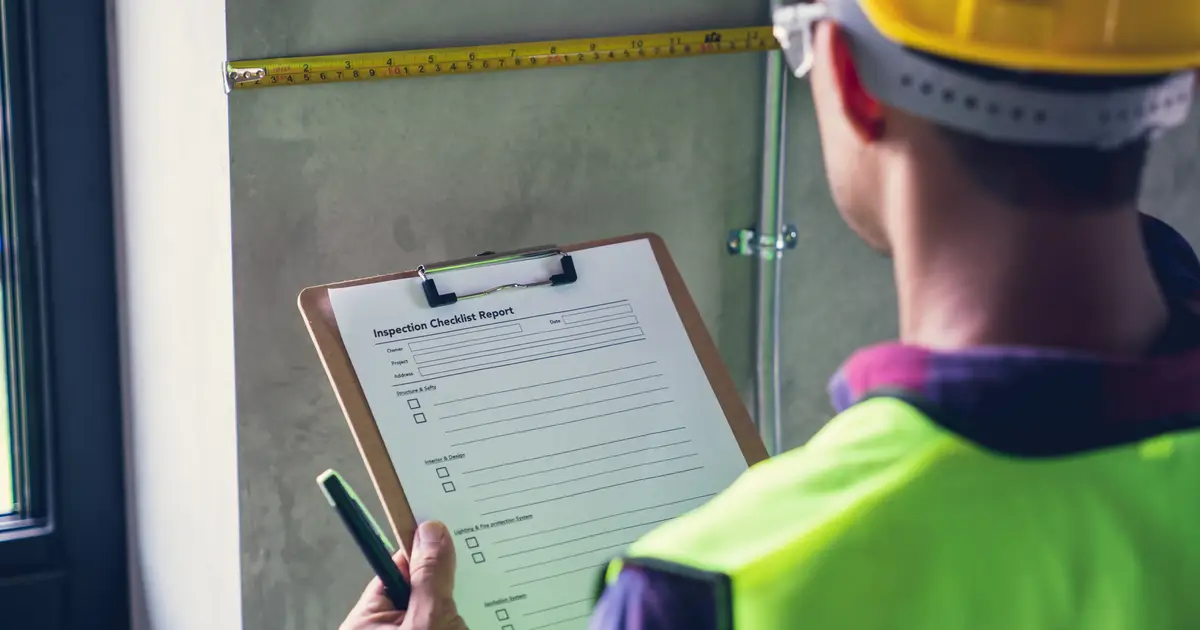 A home inspector wearing a yellow hard hat and high-visibility vest holds a clipboard with an inspection checklist while measuring a wall.