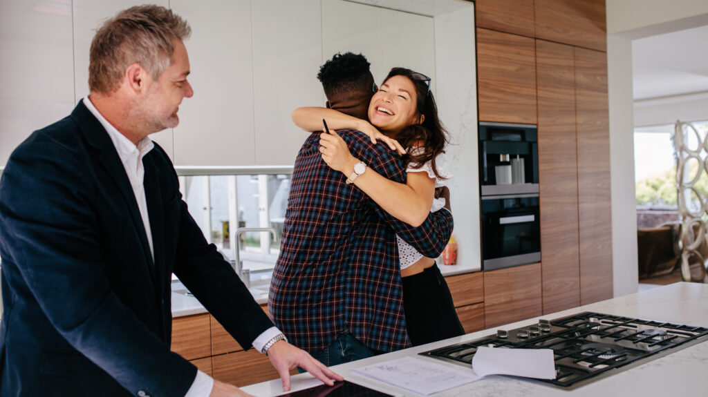 Excited couple hugging with real estate agent in kitchen of new home. Happy new property owners with realtor.