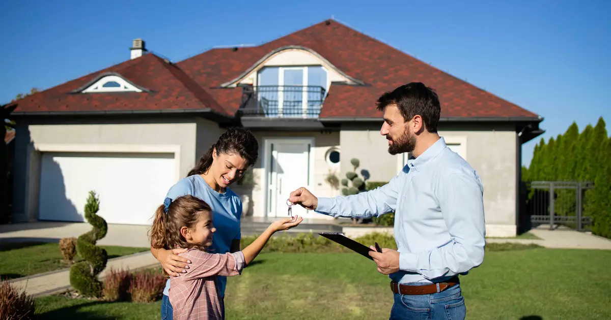 A real estate agent hands house keys to a smiling young girl while her mother embraces her in front of their new home on a sunny day.