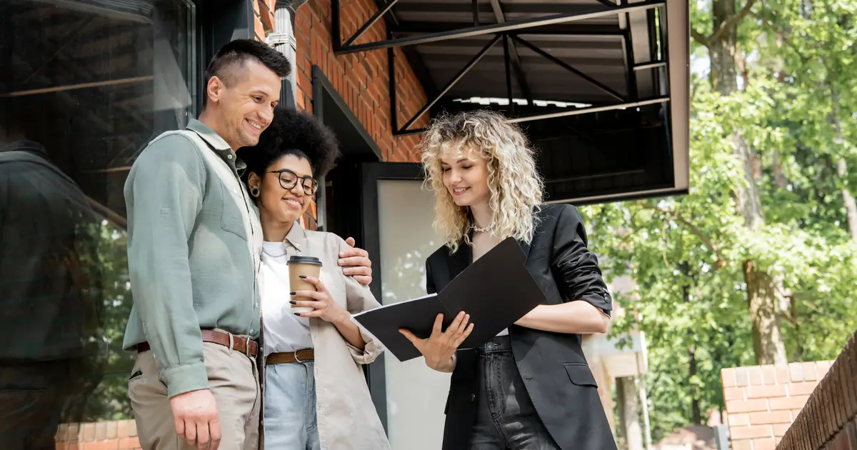 A happy couple stands outside a brick home, embracing while holding a coffee cup, as a smiling real estate agent in a black blazer shows them a folder with documents.