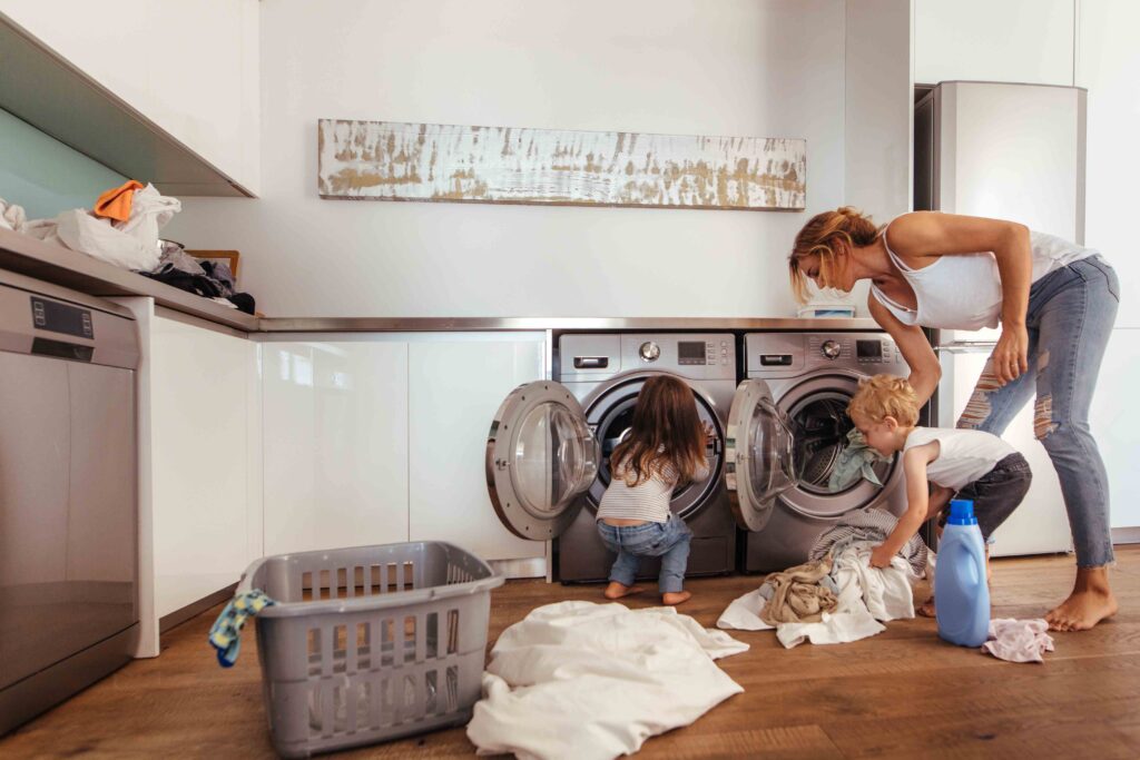 Woman with kids load clothes in washing machine. Mother and children putting laundry into washing machine at home.