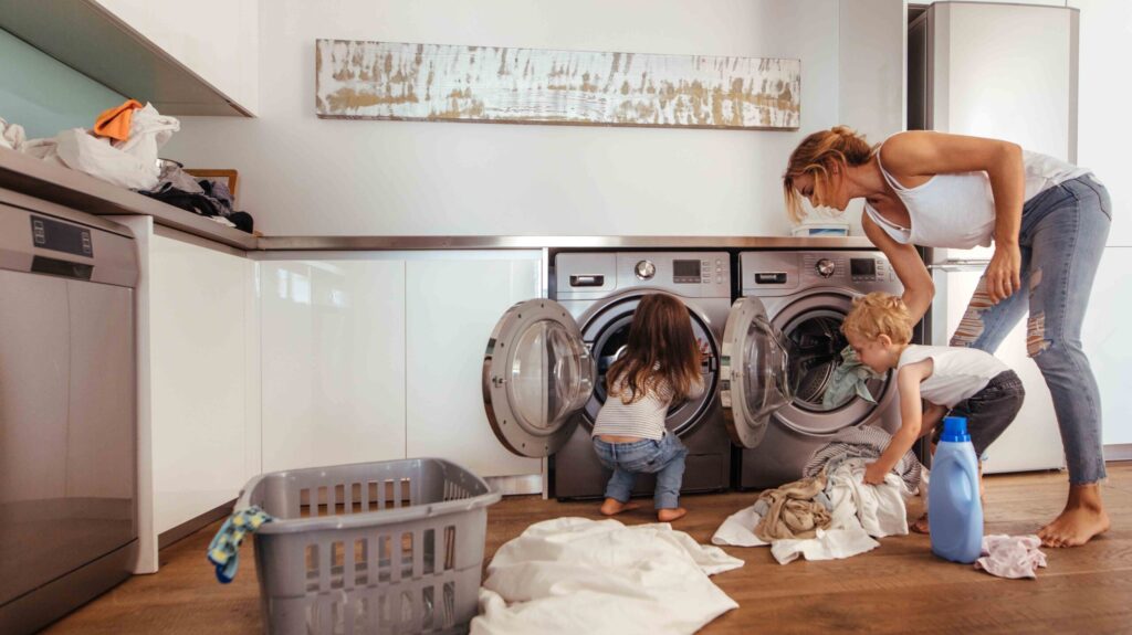 Woman with kids load clothes in washing machine. Mother and children putting laundry into washing machine at home.