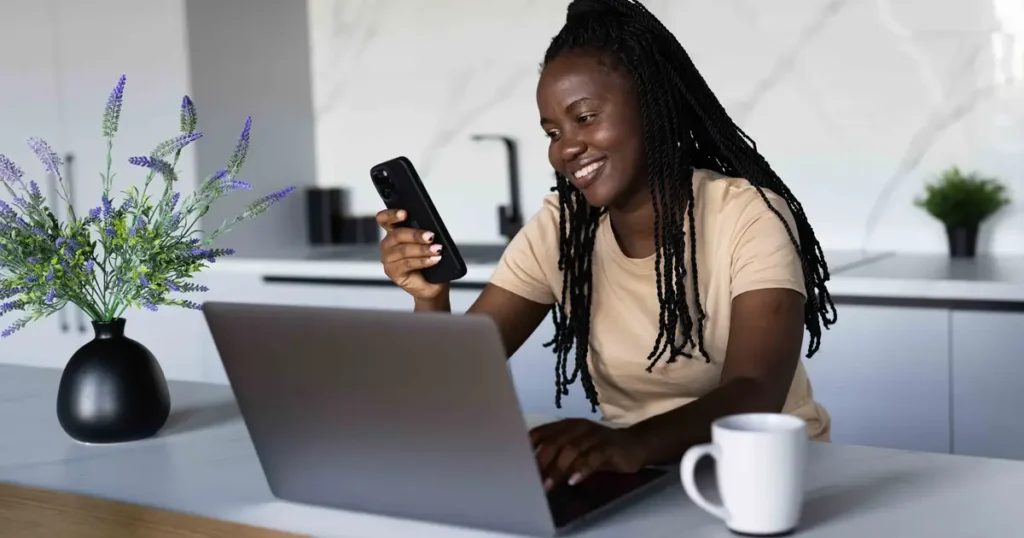 A smiling woman sitting at a kitchen counter, using her laptop and holding a smartphone, appearing engaged in a financial or mortgage-related task.