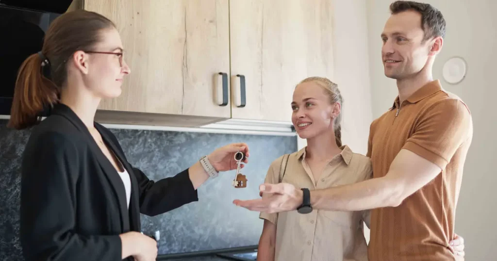 A real estate agent hands over house keys to a happy couple inside their new home, symbolizing the completion of a home purchase.