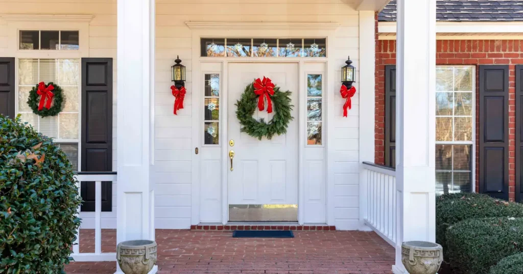 A festive front porch of a brick and white-paneled home decorated with holiday wreaths, red bows, and snowflake window decals.