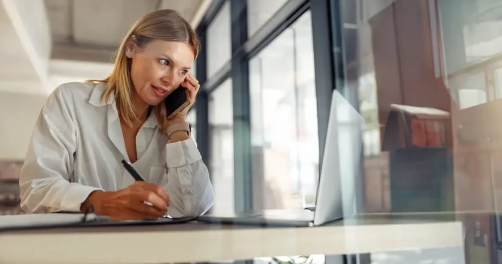 Professional woman in a white blouse talking on the phone while taking notes at her desk, working on real estate or financial planning.