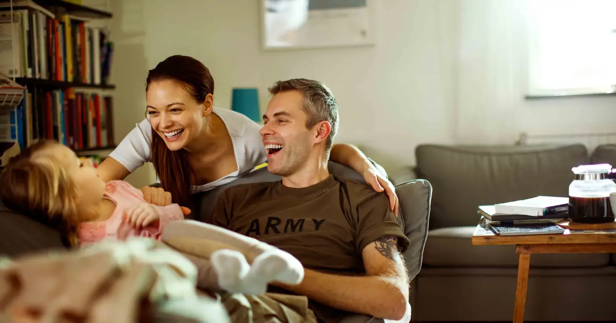 A happy military family relaxing together in their cozy living room, with a smiling mother, father in an Army shirt, and their young child.