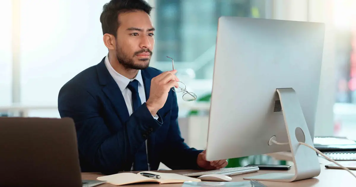 A professional man in a suit sits at a desk, holding glasses in his hand while attentively looking at a computer screen in a modern office setting.