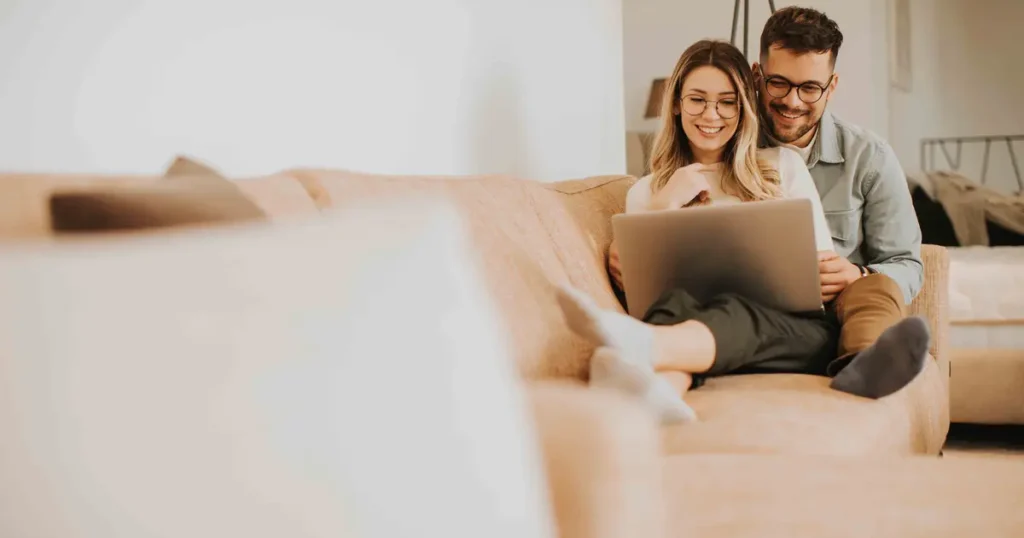 A happy couple sitting on a cozy couch, smiling while looking at a laptop, symbolizing financial planning or researching home refinancing options.