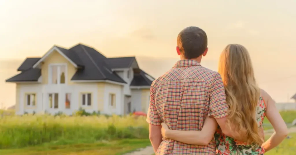 A couple standing together in a grassy field, embracing as they look at their new home in the distance, bathed in warm sunset light.