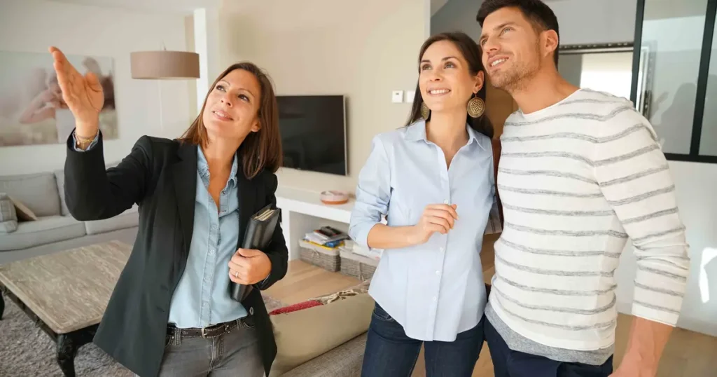 A real estate agent showing a modern home to a smiling couple, pointing out features in a bright living room as they discuss the property.