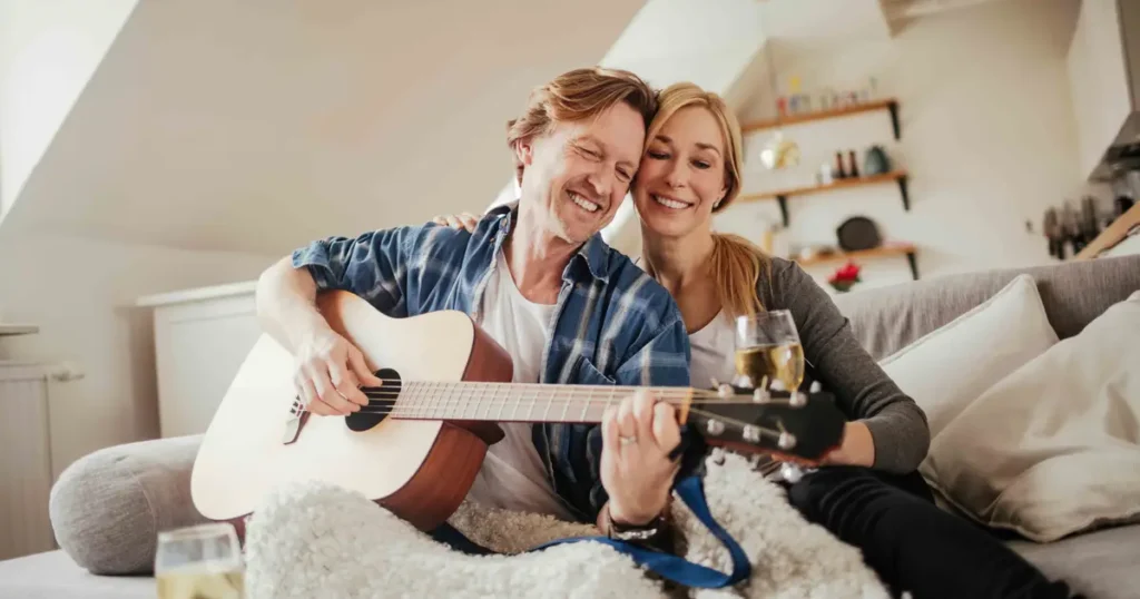 A happy couple relaxing at home on a cozy couch, with the man playing an acoustic guitar and the woman smiling while holding a glass of wine.