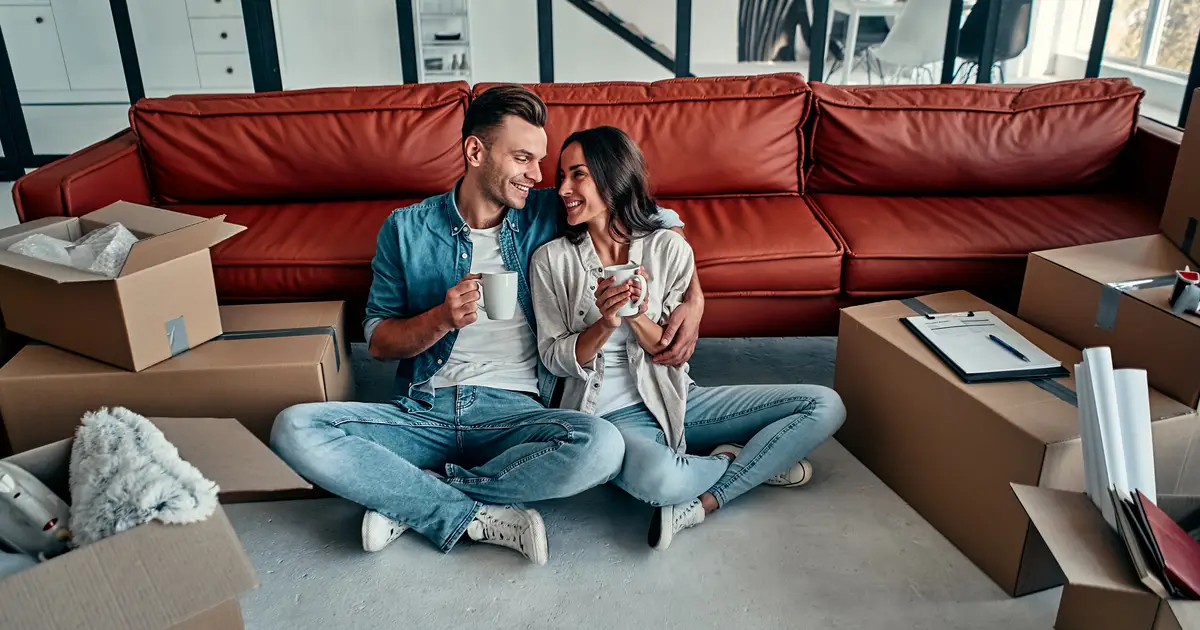 A happy couple sitting on the floor of their new home, surrounded by moving boxes, enjoying coffee and smiling at each other in a modern living space.