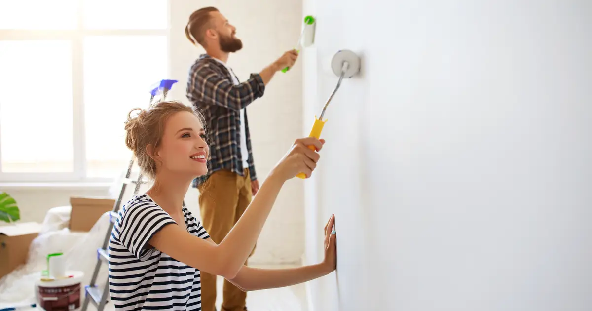 A young couple painting a bright, sunlit room together. The woman in the foreground, wearing a striped shirt, smiles while rolling white paint on the wall. The man in the background, dressed in a plaid shirt and jeans, uses a roller to paint higher areas. The room has large windows allowing natural light, with painting supplies and moving boxes scattered around, suggesting a home renovation project.