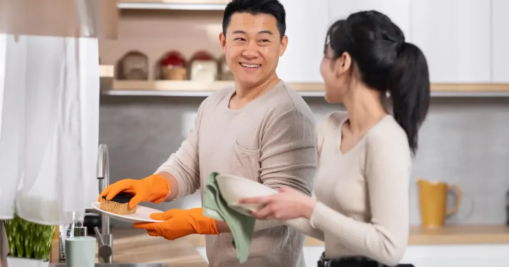 Couple smiling at each other while doing dishes in a modern kitchen; the man wears orange gloves and holds a sponge, while the woman dries dishes with a cloth