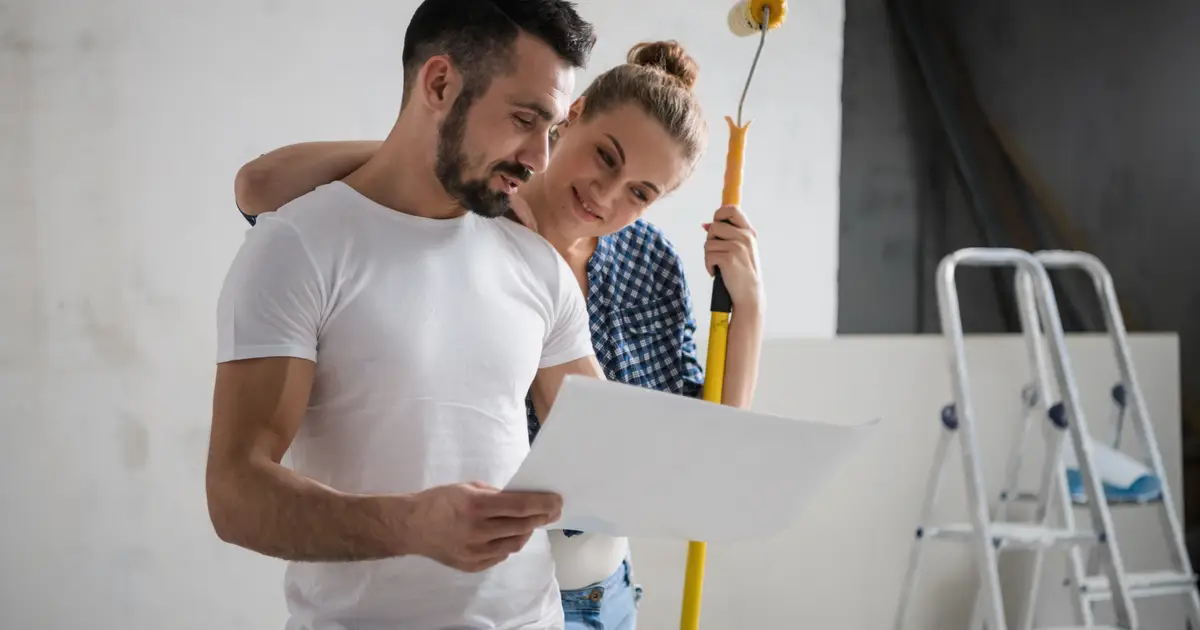 Couple reviewing renovation plans in their home, with a paint roller and ladder in the background, symbolizing a home improvement project.