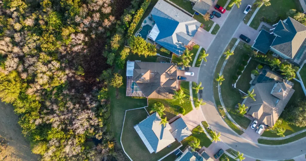 Aerial view of a suburban neighborhood with curved streets, single-family homes, and green lawns, adjacent to a wooded area with dense trees.