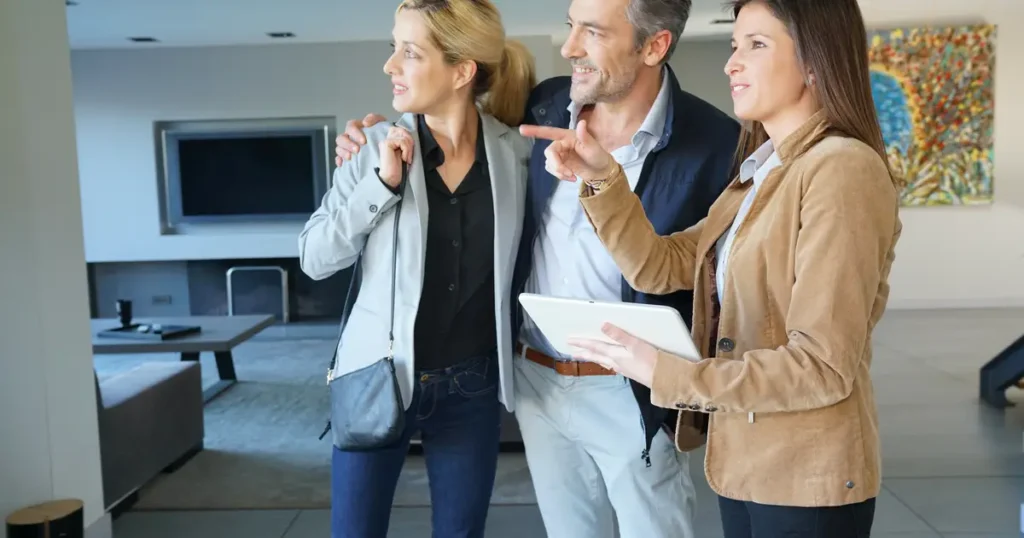 A real estate agent showing a modern home to a smiling couple, discussing features while holding a tablet in a stylish, well-lit living room.