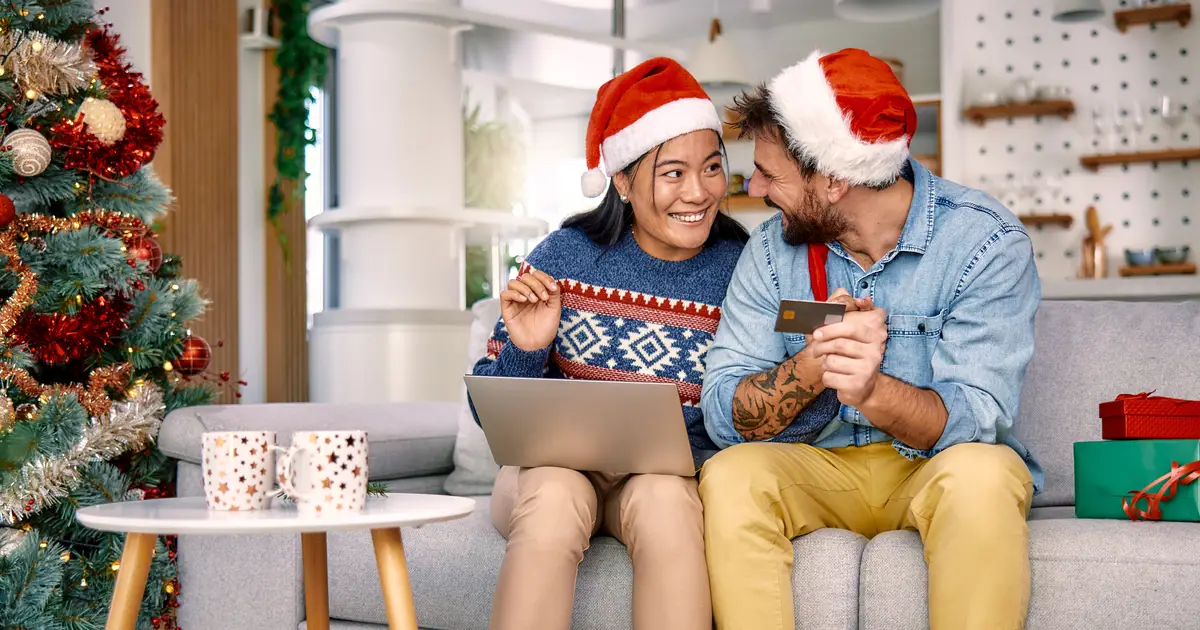 Couple wearing Santa hats sitting on a couch by a decorated Christmas tree, smiling at each other while shopping online using a laptop and credit card, with wrapped gifts and festive mugs on the table nearby.