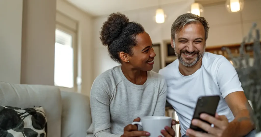 A happy couple sitting on a couch at home, smiling and looking at a smartphone together. One holds a coffee mug, creating a cozy and relaxed atmosphere.