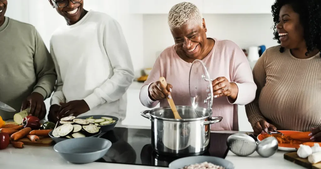 A joyful family cooking together in a bright kitchen. An elderly woman stirs a pot on the stove while two younger adults smile and prepare vegetables, including eggplants, carrots, and peppers, on a counter nearby."