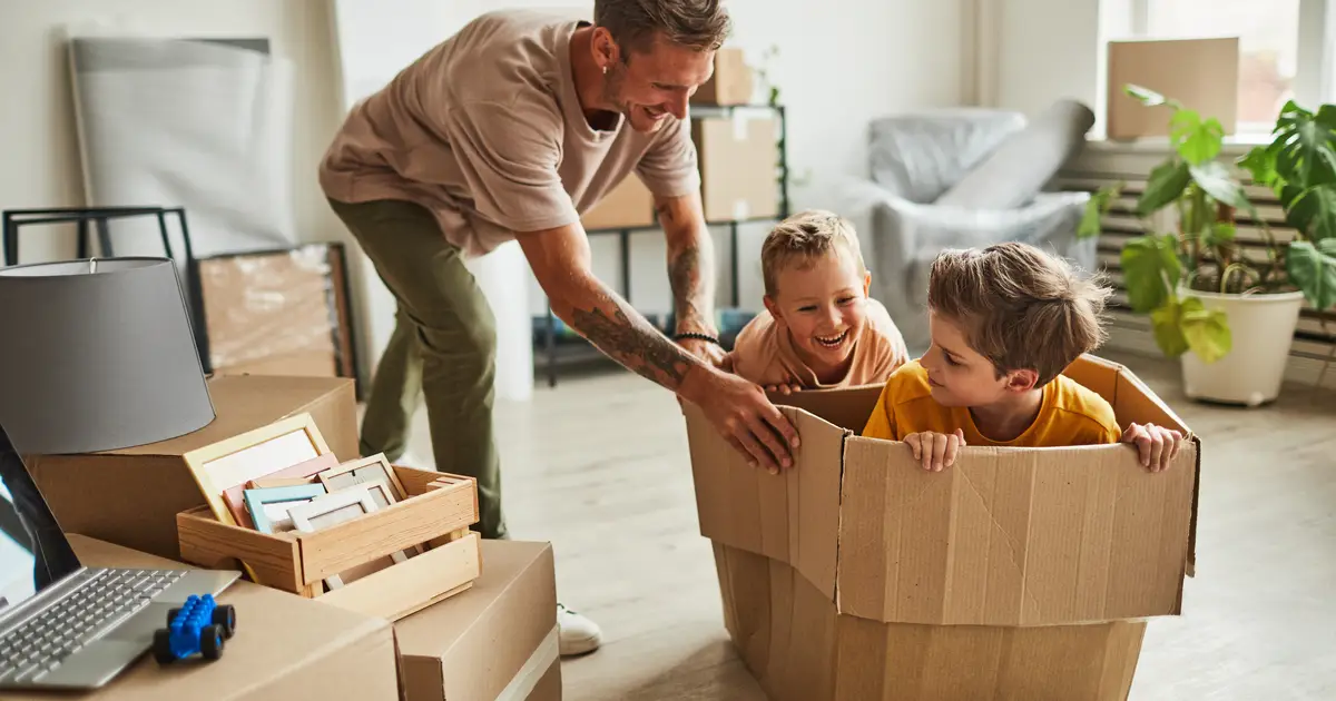 A father smiles as he plays with his two young sons, who are sitting in a large cardboard box in a living room filled with moving boxes and furniture. The scene captures a joyful moment during a home move."