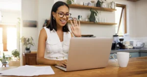 A woman wearing glasses and a sleeveless white top sits at a wooden table in a kitchen, smiling and waving at her laptop screen during a video call. Papers and a white coffee mug are on the table, with plants and kitchenware visible in the background.