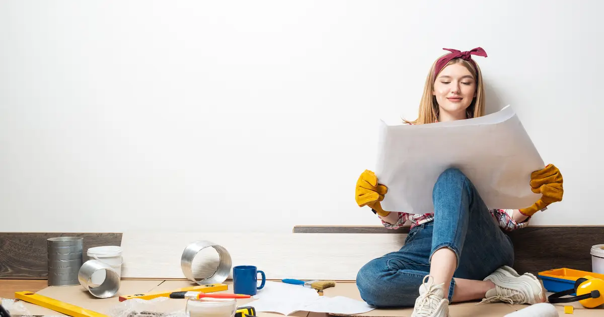 A young woman in work gloves and a headband sits on the floor, smiling while reviewing blueprints, surrounded by home renovation tools.