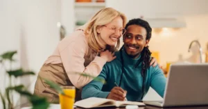 A cheerful couple working together at a kitchen table, with one person writing in a notebook and the other leaning affectionately over their shoulder. A laptop and glass of orange juice are on the table, and the background shows a bright, modern kitchen.