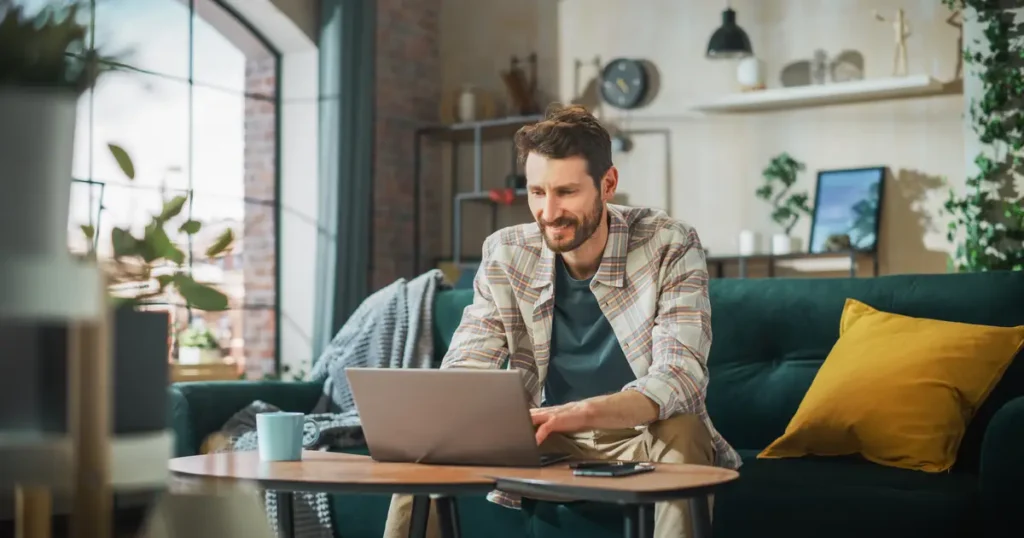 A man sitting on a green sofa in a cozy, modern living room, using a laptop on a wooden coffee table. The room features a large window, indoor plants, and minimalist decor with warm tones.